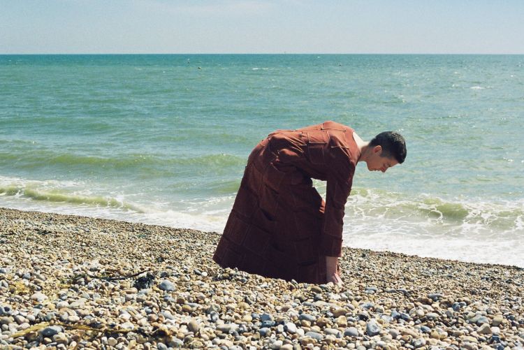 A white woman in a long red-brown dress by the sea, bending forward