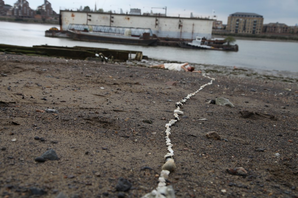 The Thames foreshore at low tide, showing a gravelly beach bisected by a long line of white stones, with a mooring block in the water. A woman in a white dress lies near the water's edge